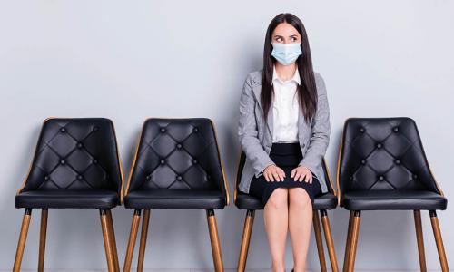 young woman wearing a mask and business attire sits alone in row of black leather office chairs while looking off to the side anxiously