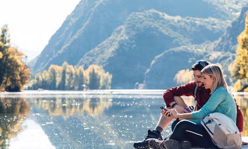 happy couple using smartphone on wooden dock above lake in front of mountains