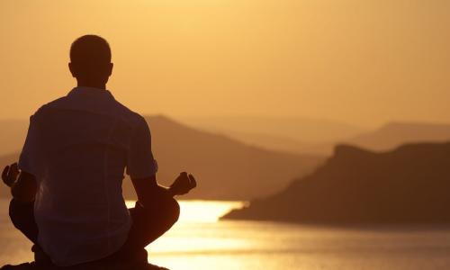 man in white collared shirt sits on rock in yoga position facing water and sunset