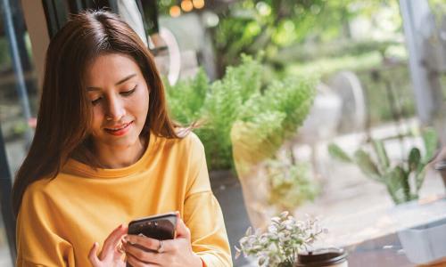 young woman in yellow shirt using smartphone in a café with lots of plants