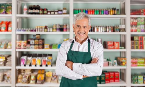 smiling silver haired shop owner in green apron stands in front of store shelves stocked with colorful items