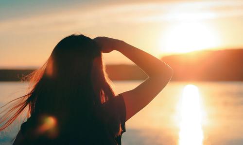 woman shields eyes while looking out over water at sun over horizon 