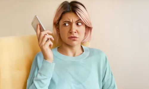 Portrait of frowning young woman in blue sweatshirt holding mobile phone