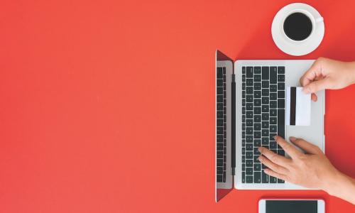 hand holding out credit card while typing information into laptop next to coffee cup and smartphone on red background 