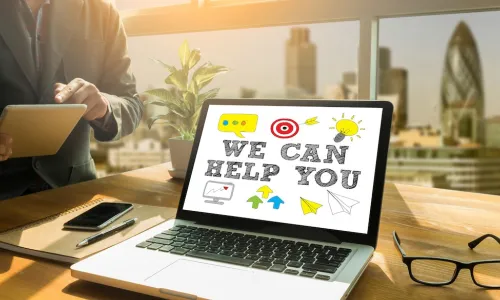 young businessman works on tablet while laptop on desk displays message "We Can Help You"