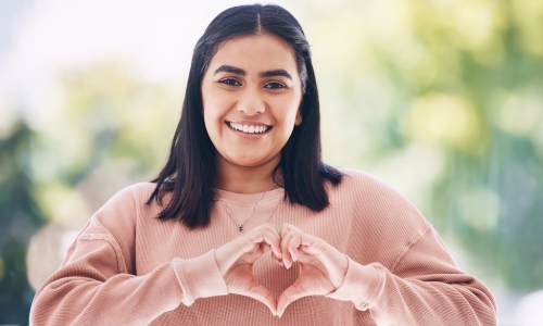 young Gen Z woman in pink sweatshirt making a heart shape with her hands
