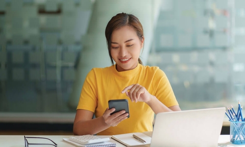 young woman uses online banking on her phone while reviewing financial reports