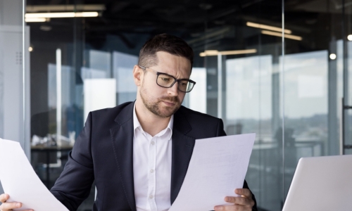 a businessman at a desk reviews with a perplexed expression on his face reviews his executive benefits plan on various papers