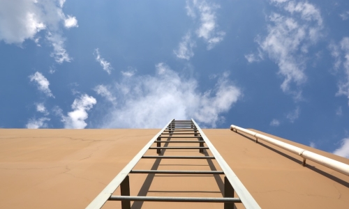 ladder leaning against biege stucco wall leading up to blue sky