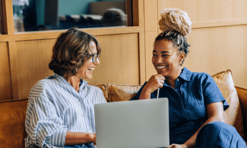 two women smiling sitting on a couch looking at a laptop
